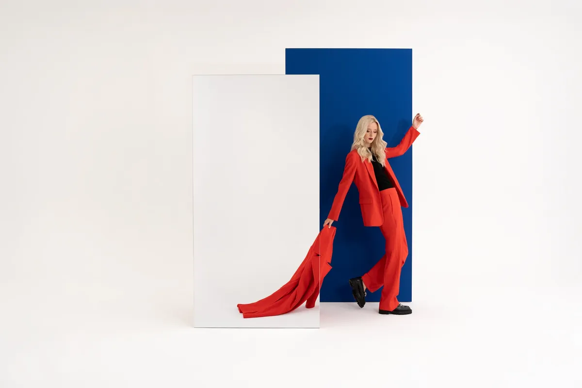 AppelrathCüpper, blondes Model im roten Hosenanzug zieht einen roten Mantel hinter sich her, blaue Fläche und Spiegel, Kampagne 2022, fotografiert von Kilian Amrehn