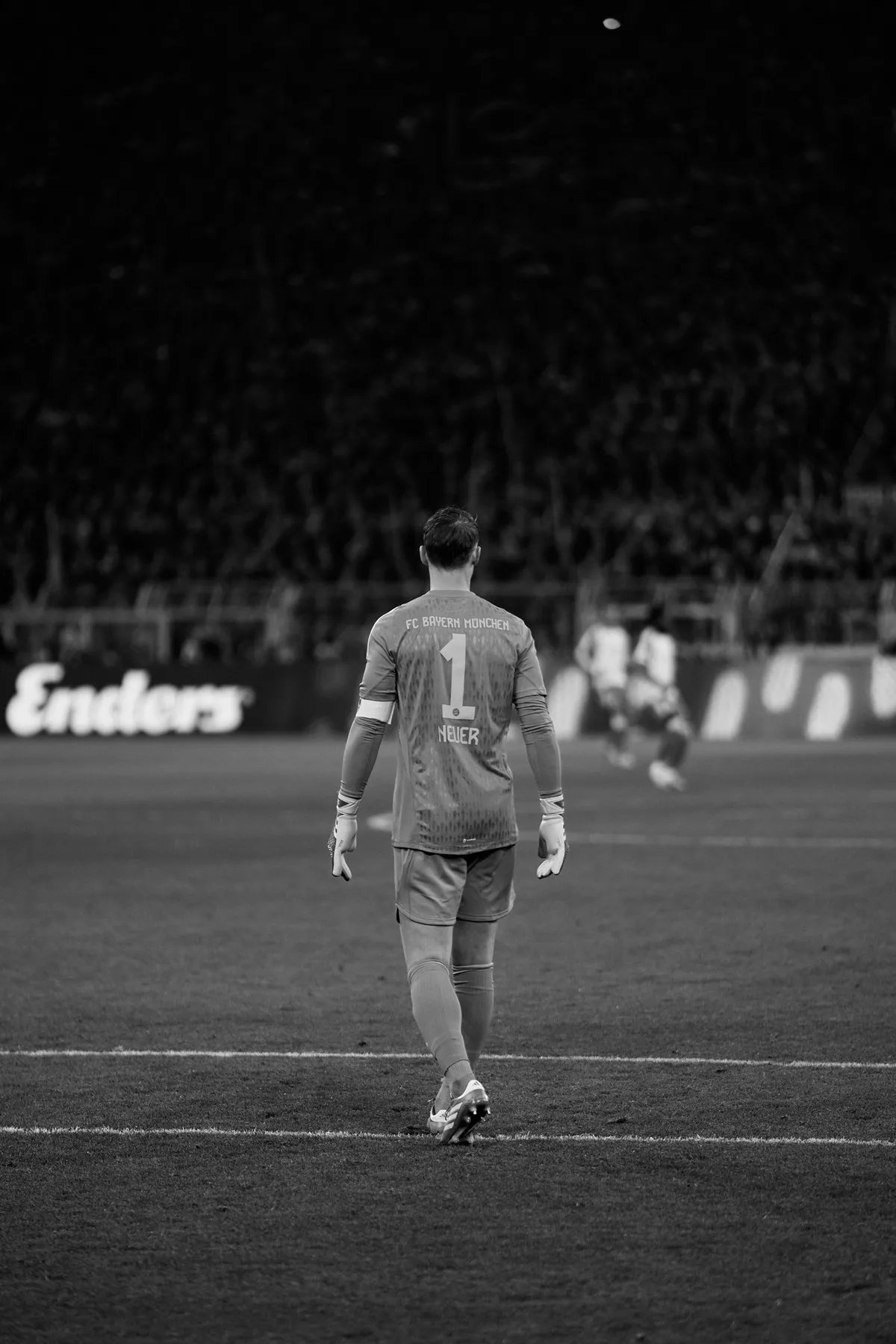 Bundesliga, Torwart mit der Nummer 1 geht allein über den Rasen vor dunkler Tribüne, Rückansicht in Schwarzweiß, Sportfotografie, fotografiert von Kilian Amrehn