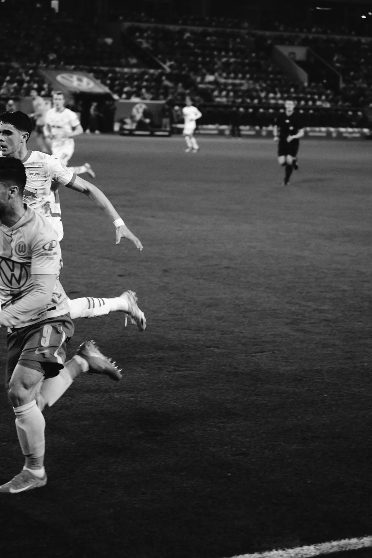 Bundesliga, Spieler im Sprint dicht am Bildrand, Schiedsrichter im Hintergrund, Schwarzweiß im Flutlicht, Sportfotografie, fotografiert von Kilian Amrehn