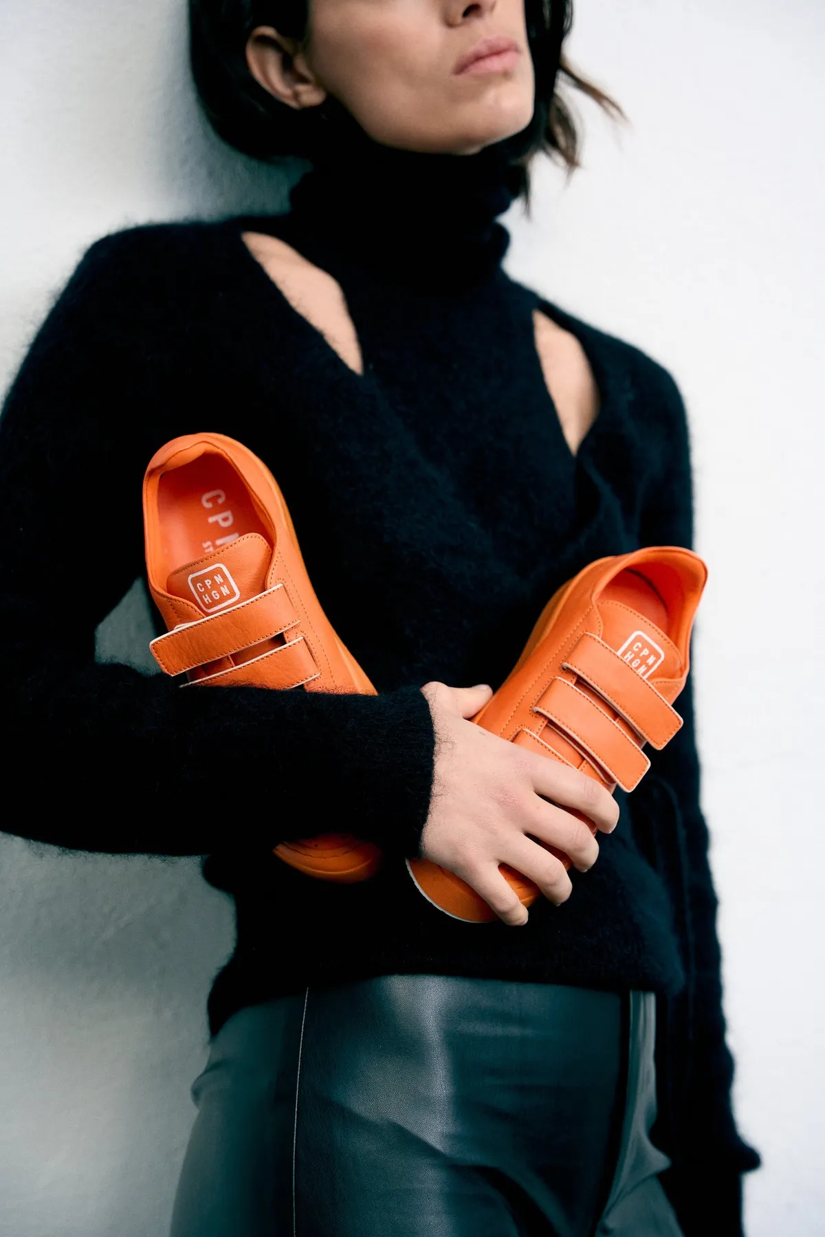 Copenhagen Studios, Model im schwarzen Cut-out-Pullover hält orangefarbene Klettverschluss-Sneaker im Arm, Lederhose, Kampagne Oktober 2022, fotografiert von Kilian Amrehn