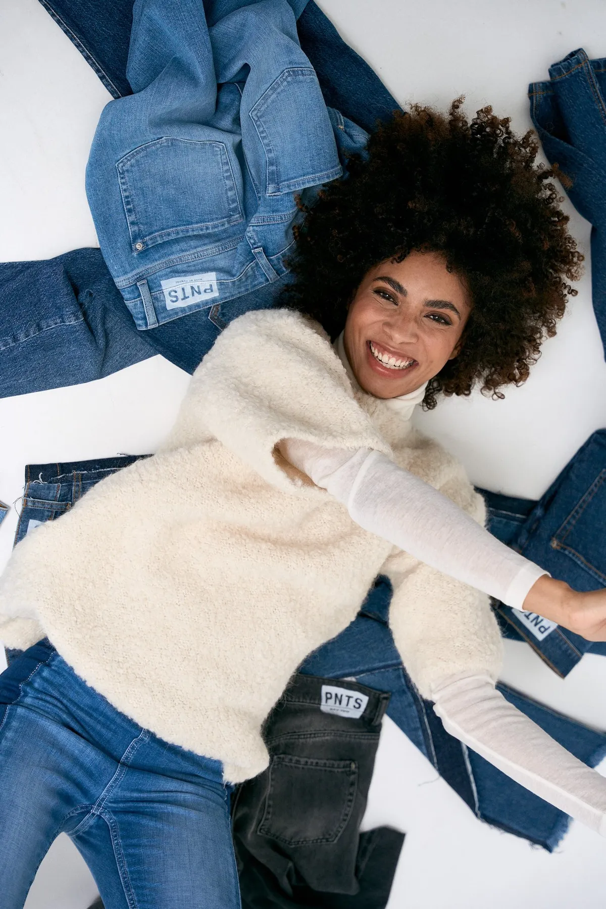 PNTS, lachendes Model mit Afro im flauschigen Pullunder liegt auf ausgebreiteten Jeans mit PNTS-Patches, Kampagne 2022, fotografiert von Kilian Amrehn