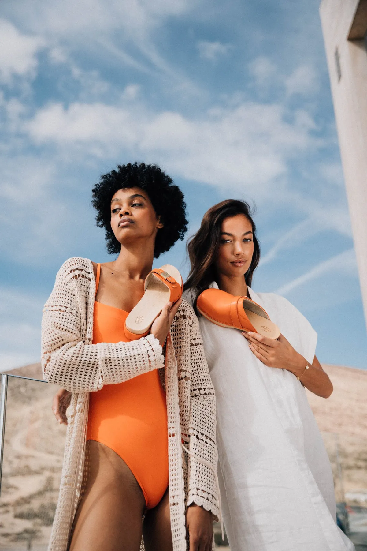 Scholl iconic, zwei Models in Badeanzug und Strickjacke halten orangefarbene Pantoletten vor blauem Himmel, Kampagne Fuerteventura 2021, fotografiert von Kilian Amrehn