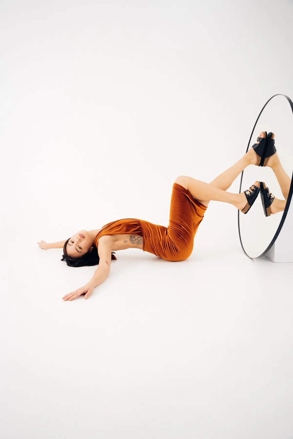 Scholl iconic, Model im rostroten Kleid liegt am Boden, Füße mit schwarzen Sandalen spiegeln sich im runden Spiegel, Kampagne Oktober 2021, fotografiert von Kilian Amrehn