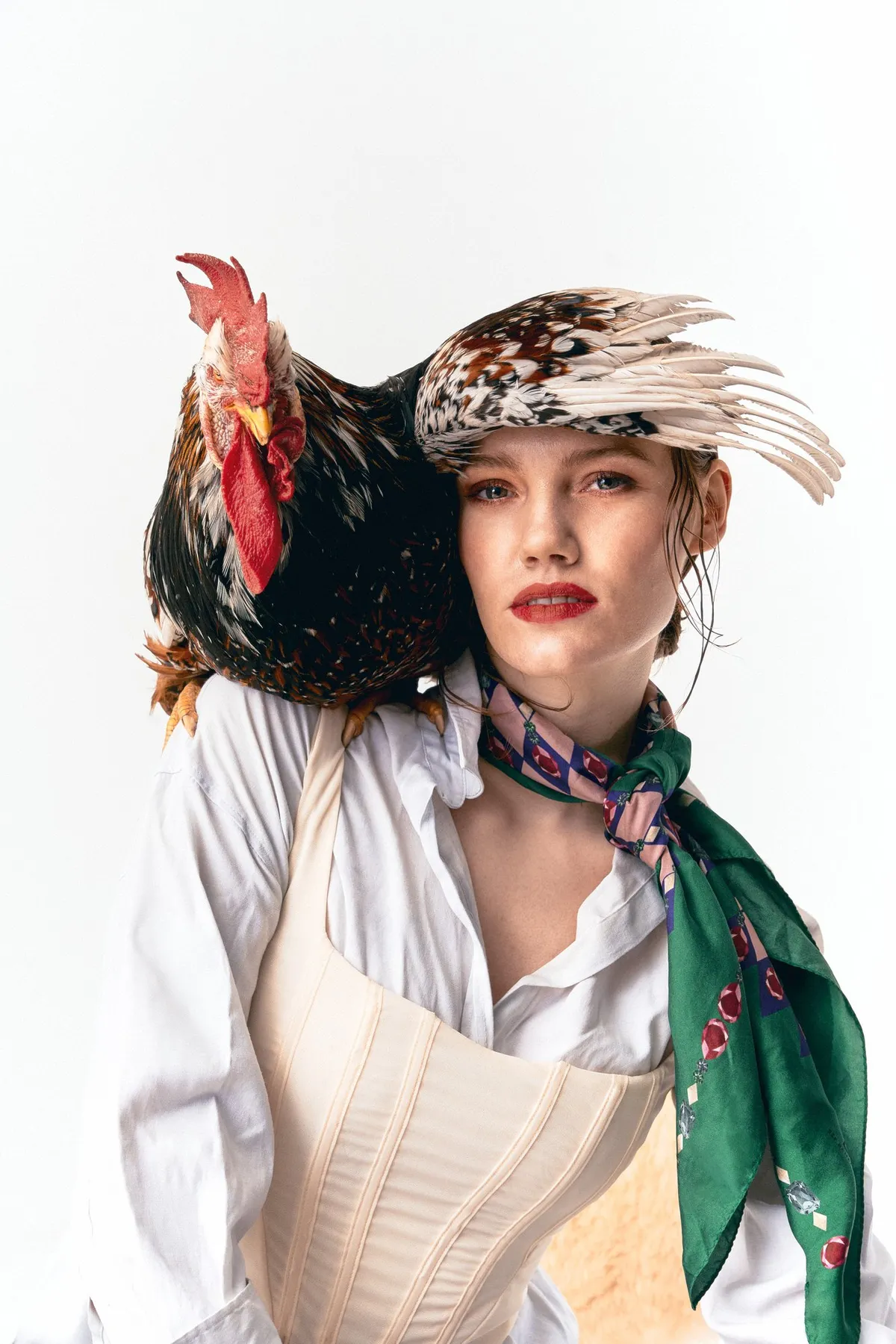Untamed, Model mit Federkappe, Korsage und grünem Seidentuch, ein Hahn auf der Schulter, roter Lippenstift vor Weiß, Editorial 2024, fotografiert von Kilian Amrehn